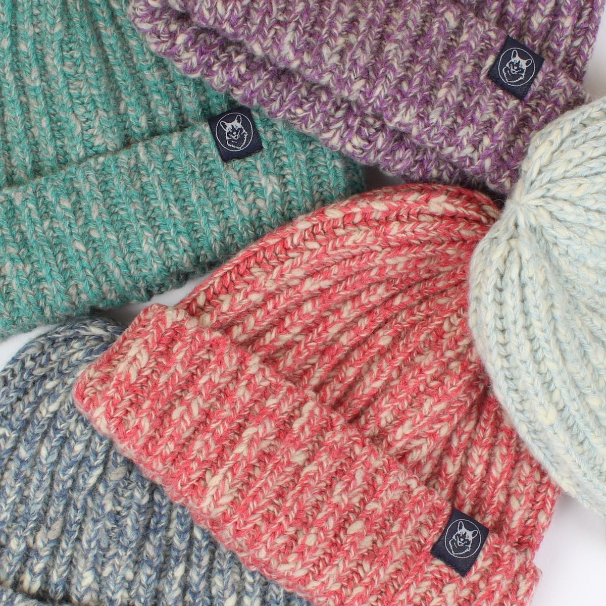 Four marled knit beanie hats arranged in a flat lay, featuring a teal, purple, coral pink, and heather blue colourway. Each beanie has a small black square brand patch on the cuff with a white corgi dog logo.