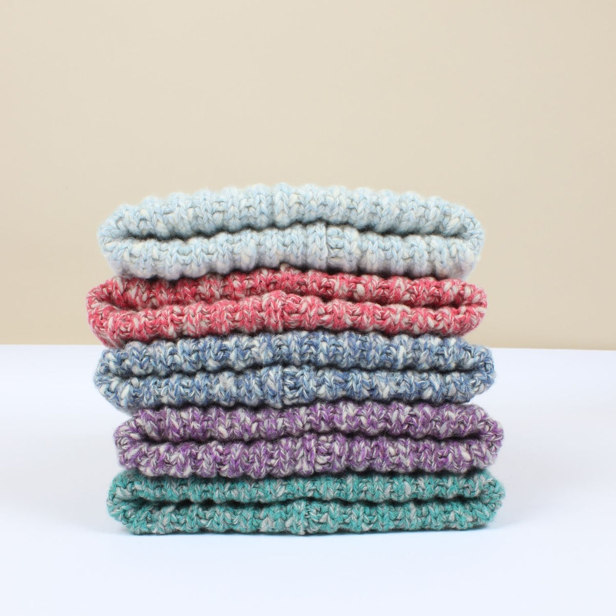 A stack of six folded textured beanies in pastel and heathered colours, including light blue, coral pink, medium blue, lavender purple, and teal green, set against a neutral beige and white background.