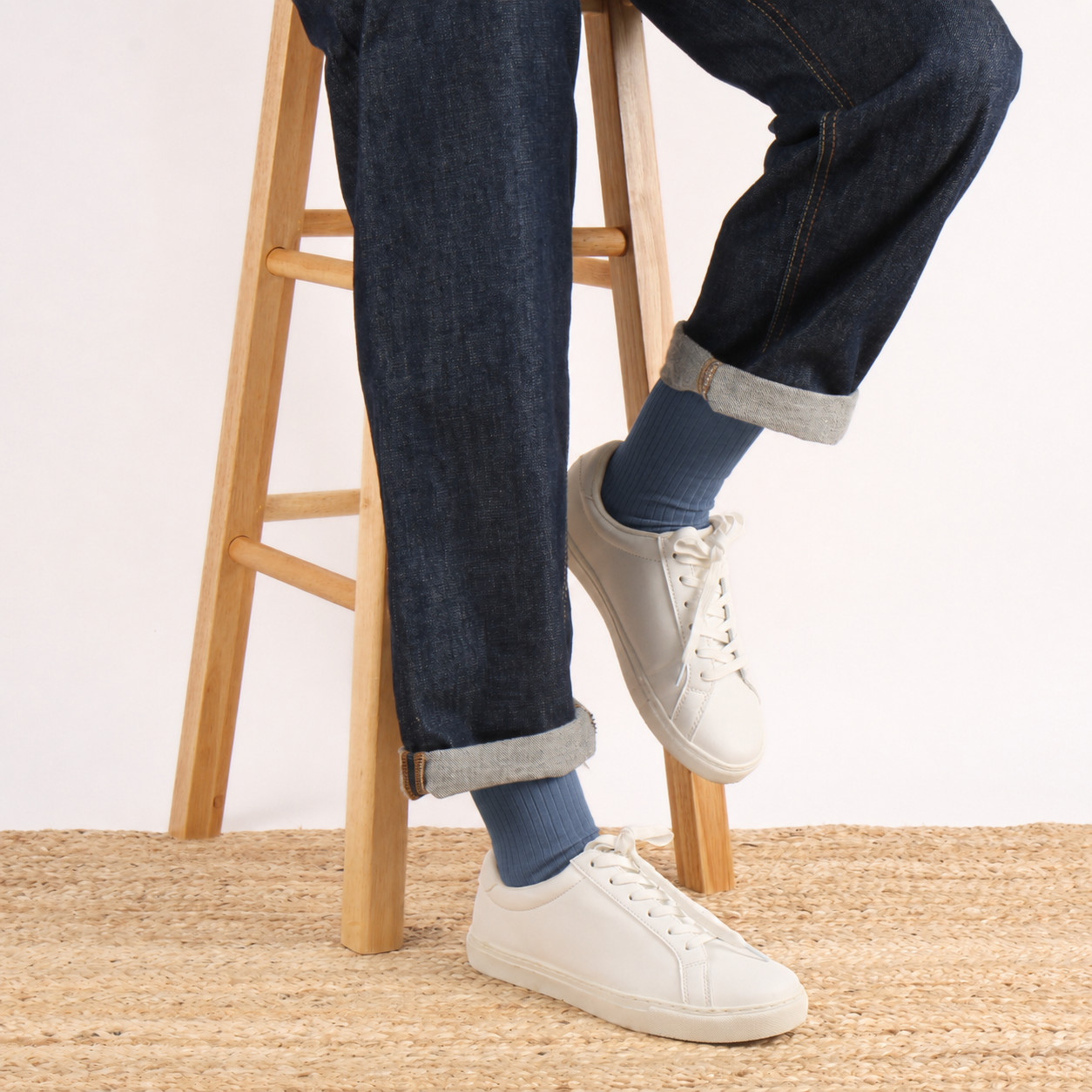 A person seated on a wooden stool wearing dark blue jeans with rolled cuffs, blue socks, and white low-top sneakers, photographed against a plain light background.