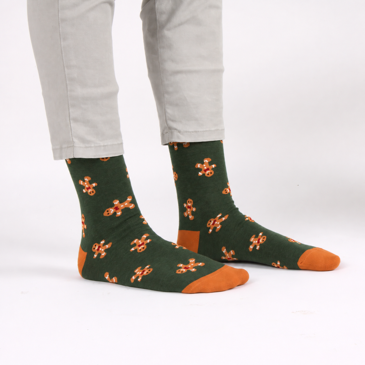 A person wearing dark green crew socks with an orange gingerbread man pattern and solid orange toes, heels, and soles, paired with light gray pants on a white studio background.