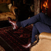 Person wearing brown leather shoes and black socks sitting in a cosy room with a fireplace.