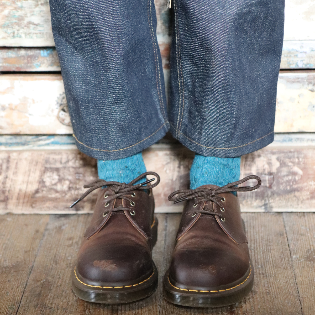 A close-up of a person's feet wearing dark brown leather Dr. Martens lace-up shoes, blue denim jeans, and bright blue socks, standing on a rustic wooden floor against a weathered wood plank wall.