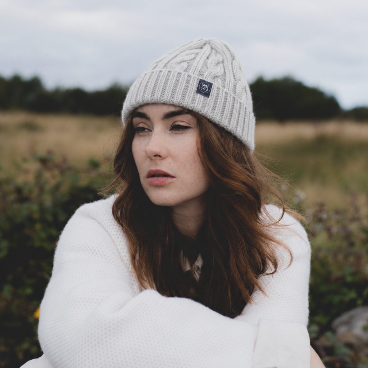 A woman wearing a light gray cable-knit beanie with a small black brand patch and a white knit sweater, photographed outdoors in a grassy field under an overcast sky.