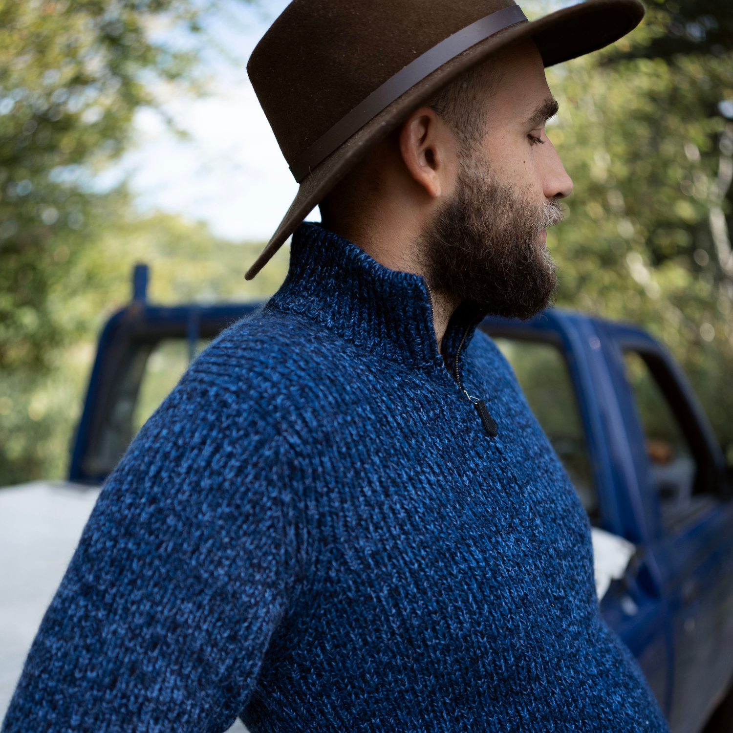 Man wearing a blue sweater and brown hat standing next to a blue truck in a forest setting