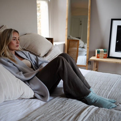 A woman sitting on a bed with pillows, wearing luxury cashmere and silk socks in a neutral color.