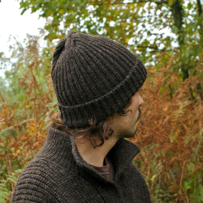 Person wearing a dark knit beanie and coat in an outdoor setting with trees and bushes.