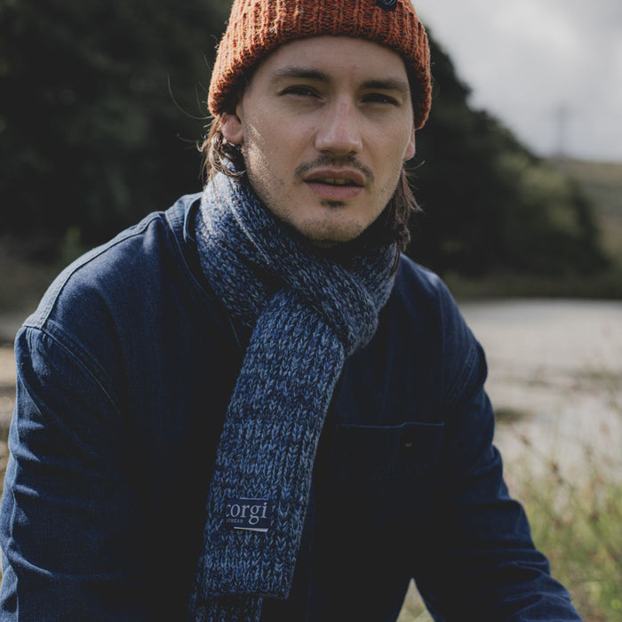 A man wearing a blue jacket and a blue marl knitted scarf with a ribbed pattern, sitting outdoors with a field in the background.