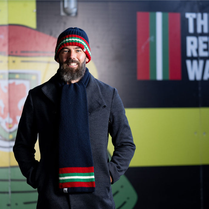 A person wearing a navy coat and a beanie hat with red and green stripes, standing in front of a wall with colorful artwork.