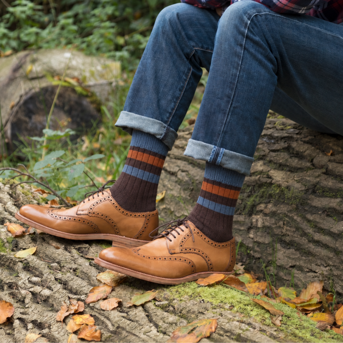 A person sitting outdoors on a log wearing tan brogue oxford shoes, blue jeans, and dark socks with orange and blue stripes. Fallen leaves and greenery are visible around the legs and shoes.