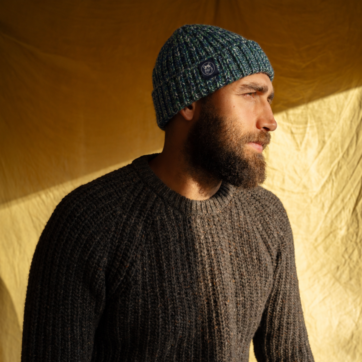 A man wearing a dark green and black knit beanie with a small circular logo patch, paired with a dark gray ribbed knit sweater, set against a warm, yellow-toned tent backdrop.