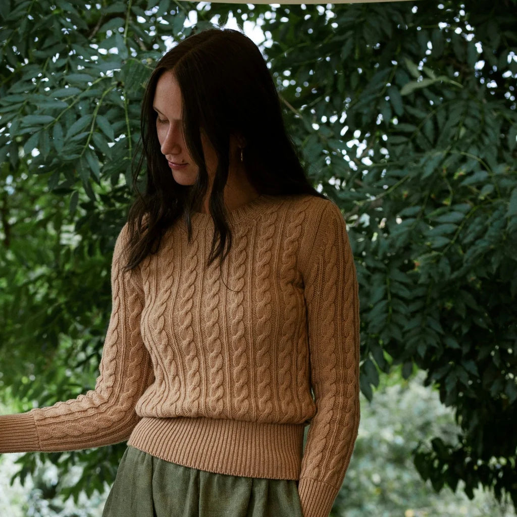A woman wearing a beige cable-knit sweater and an olive green skirt, standing outdoors in front of a background of green foliage.
