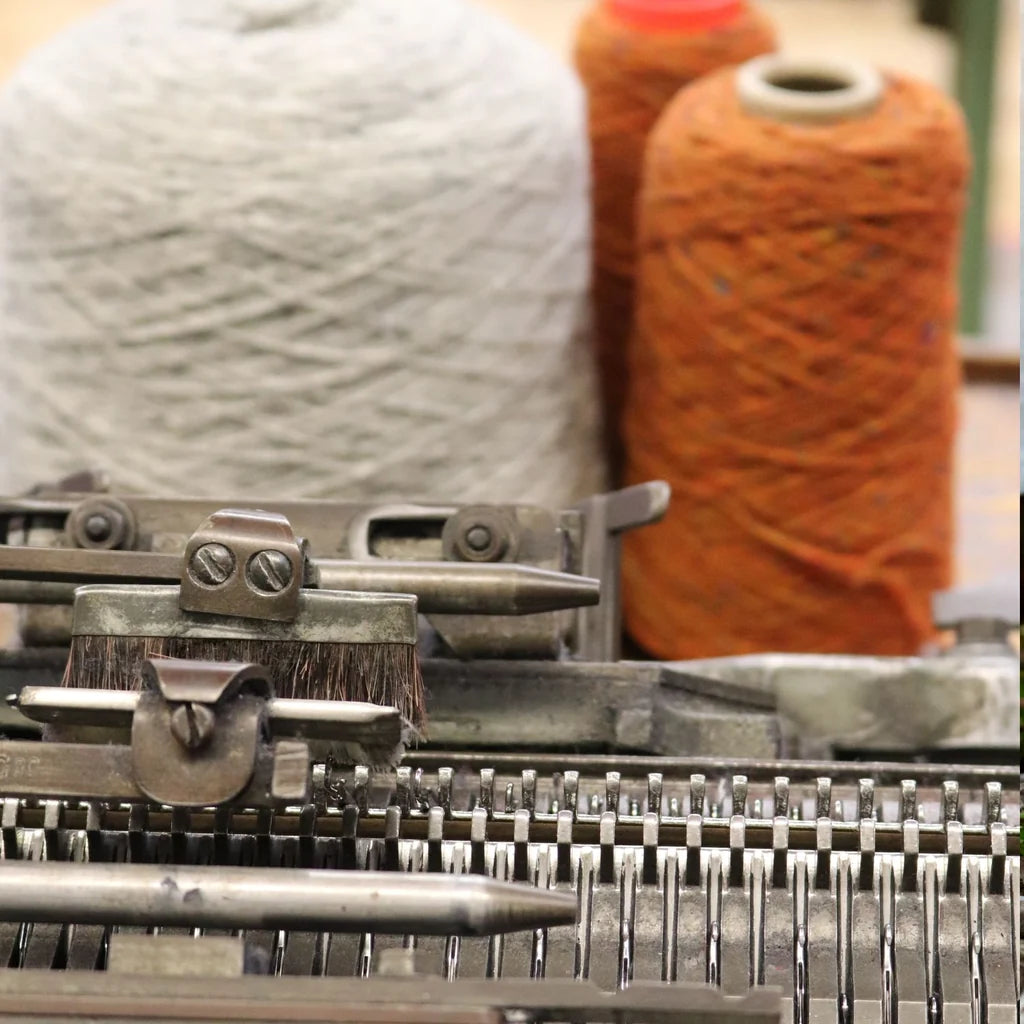 Close-up of a knitting machine with metal needles in the foreground and two large yarn cones, one white and one rust orange, blurred in the background.
