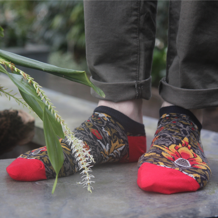 A pair of multicolored trainer socks with a geometric pattern, predominantly red and black, worn on someone's feet, with greenery in the background.