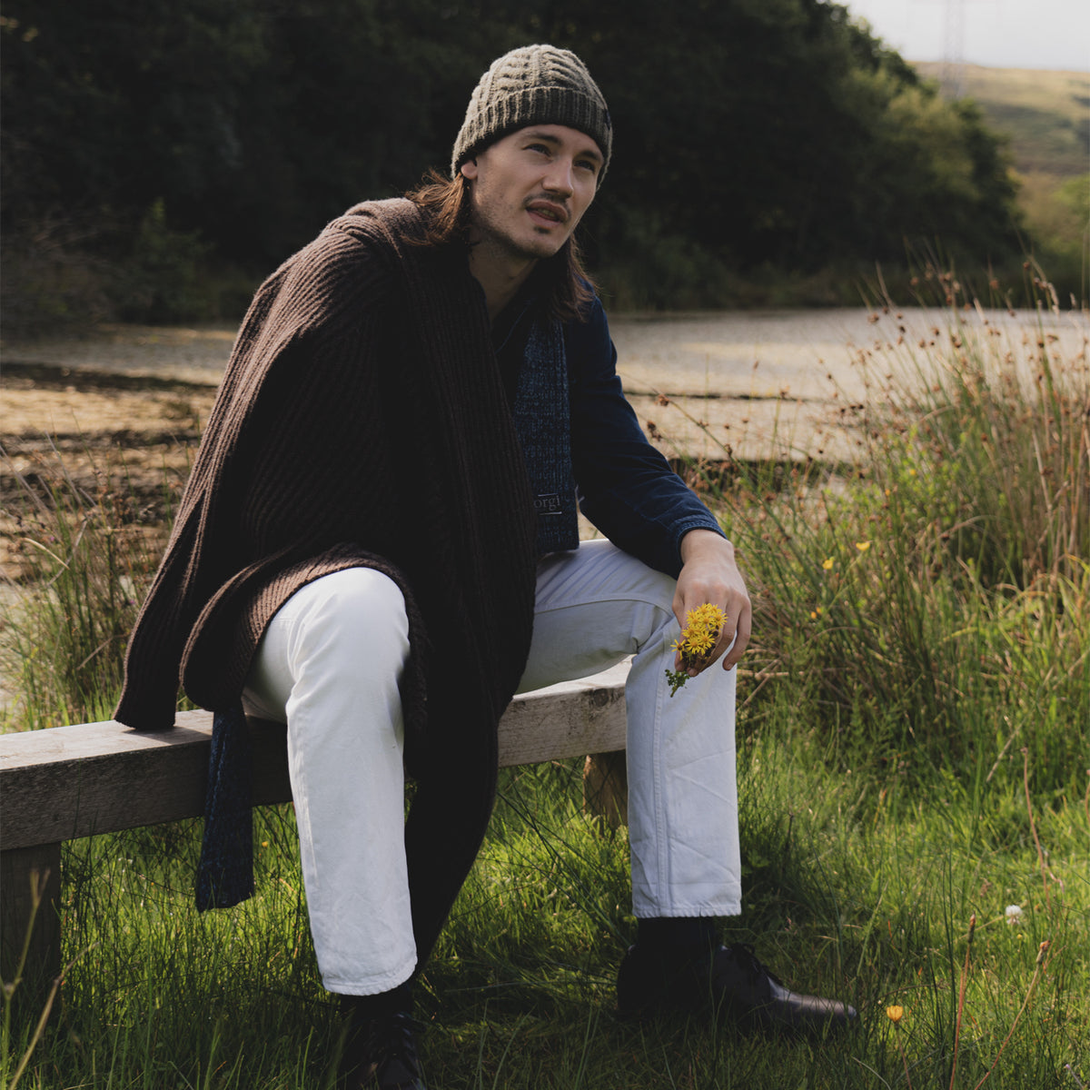 A man sits on a wooden bench outdoors in a grassy field, wearing a brown knit beanie, a dark brown ribbed cardigan over a navy shirt, white trousers, and black boots, while holding a small bunch of yellow flowers.