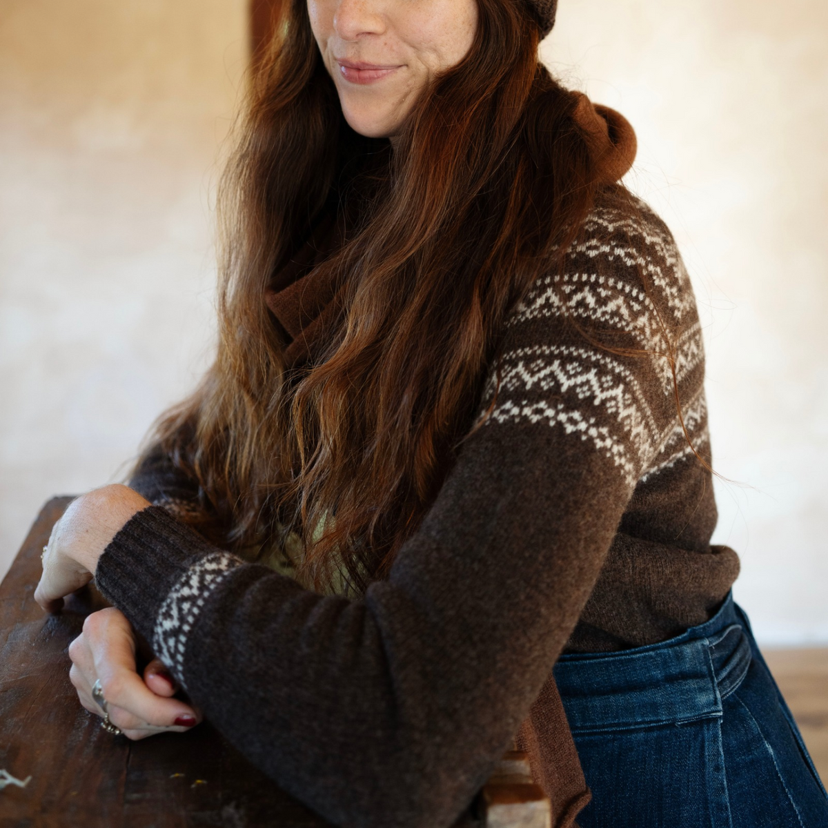 A woman wearing a dark brown knit sweater with a cream Fair Isle pattern on the yoke and cuffs, leaning over a rustic wooden table.