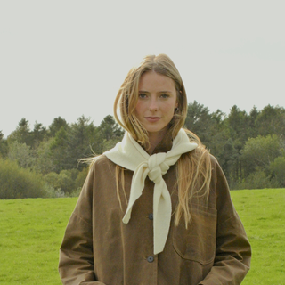 Woman wearing a brown jacket and white scarf standing in a grassy field with trees in the background