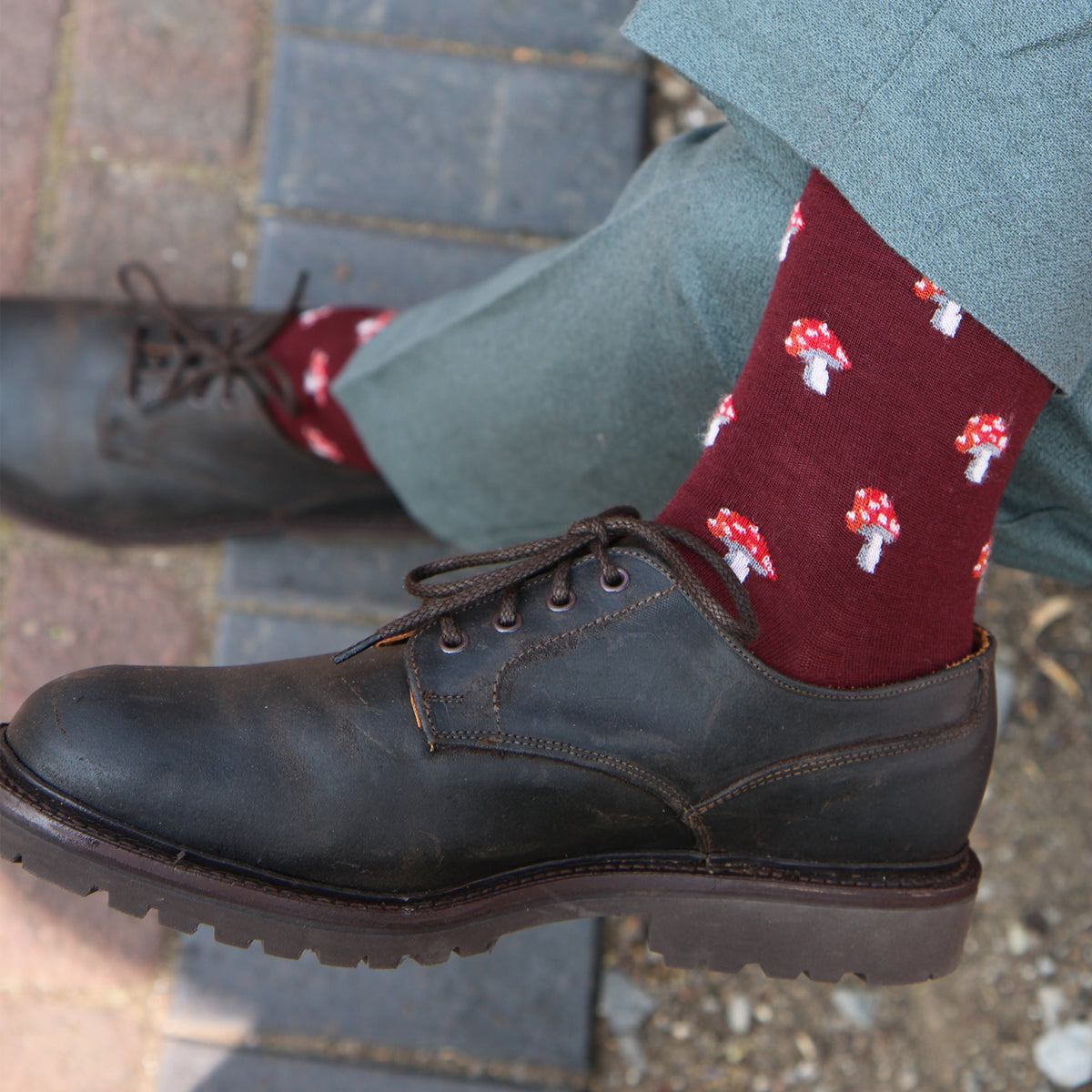A close-up of a person's feet wearing dark brown leather lace-up shoes, olive green trousers, and burgundy socks with a red and white mushroom print, standing on a brick pavement.