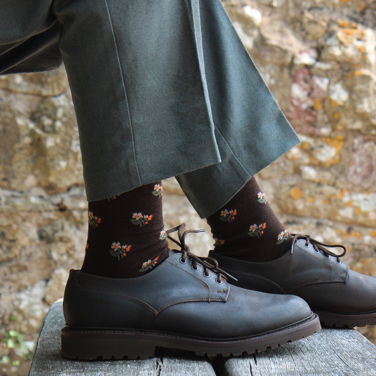 A close-up of a person's legs wearing dark brown floral socks and dark brown leather oxford shoes, standing on a weathered wooden surface with a rustic stone wall in the background.