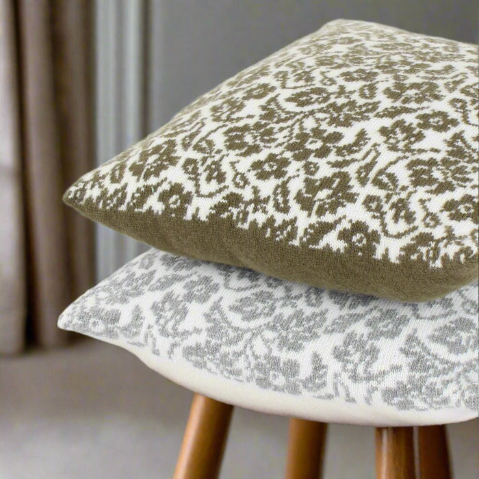 Two square floral patterned wool cushions on a wooden stool, one with a darker green floral pattern and the other with a lighter white floral pattern, both with envelope style closures.