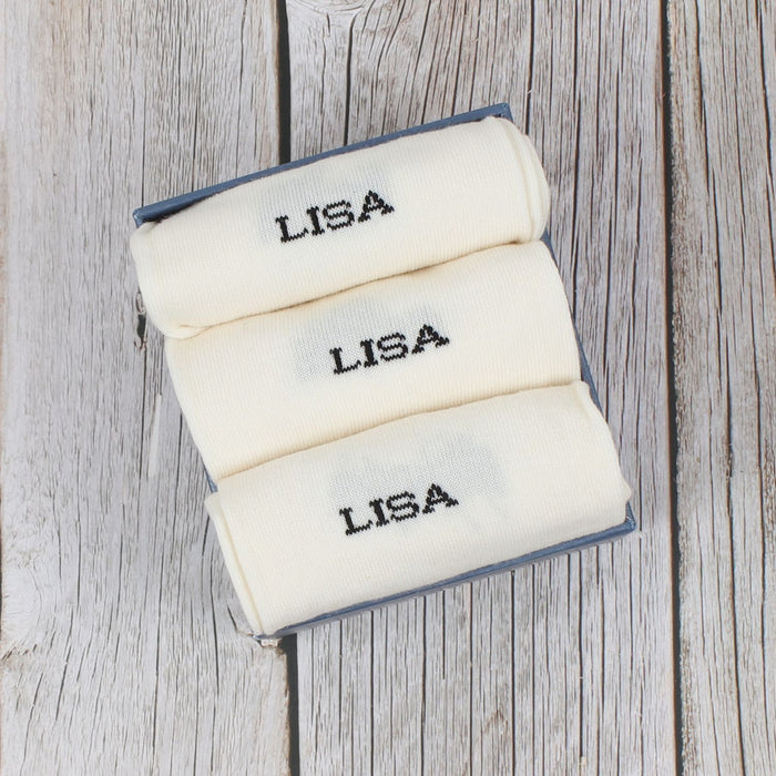 Three folded personalized socks with the name 'LISA' knitted on the side, displayed on a wooden surface.