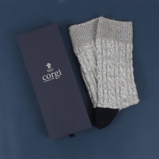 A pair of hand-knitted gray cable cashmere and cotton socks displayed alongside its packaging box with the brand 'Corgi' visible.