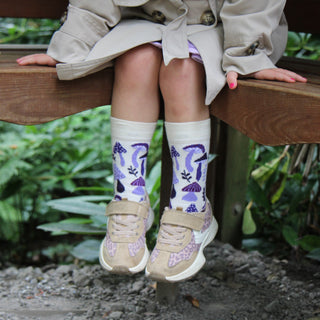 Child sitting on a wooden bench wearing beige shoes with purple patterns and white socks with purple designs.