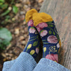 Child wearing floral socks with yellow toes and heels, sitting against a wooden post outdoors.