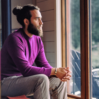 A man wearing a purple classic crew sweater with a crew neck, sitting by a window.