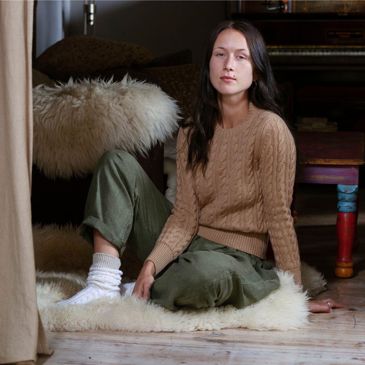 A woman sitting on the floor wearing a beige cable crew neck pullover and green pants.
