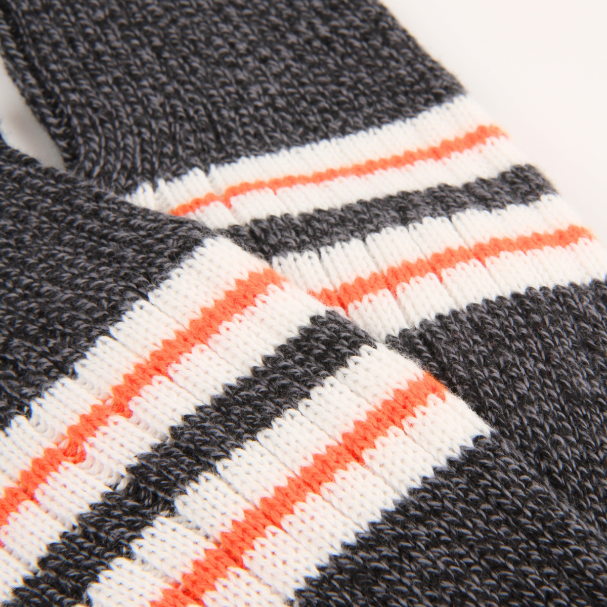 Close-up of a knitted textile with horizontal stripes in dark charcoal gray, white, and bright orange on a plain light background.