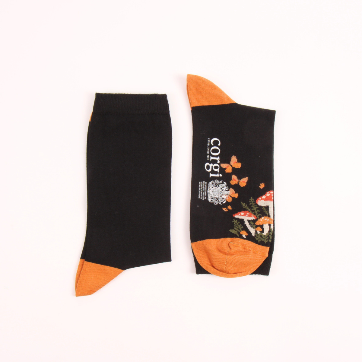A pair of black crew socks with orange tips and heels, featuring a printed design of red mushrooms, orange butterflies, and white text reading 'corgi' on one sock, laid flat on a white background.