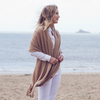 A woman standing on a beach wearing a beige cashmere wrap, white top, and white pants.