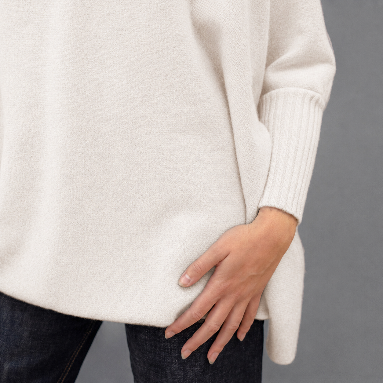 Close-up of a person wearing a soft off-white knit sweater with dark jeans, showing the sleeve cuff and hand against a plain gray background.