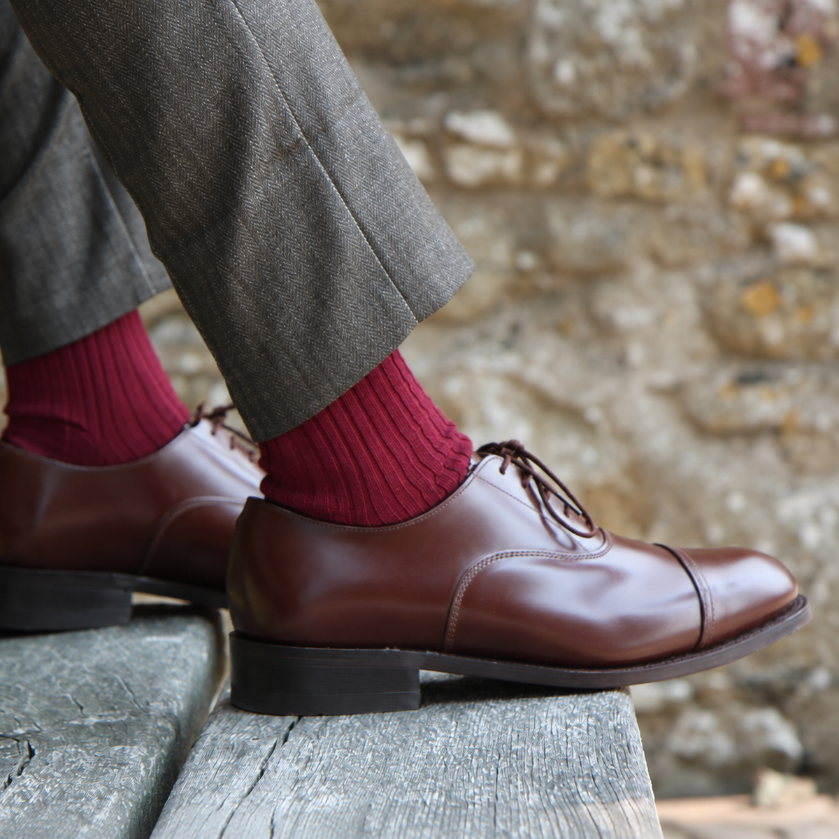 A close-up of a person's legs wearing brown leather cap-toe oxford shoes, dark gray trousers, and red ribbed socks, sitting on a weathered wooden bench with a blurred stone wall background.