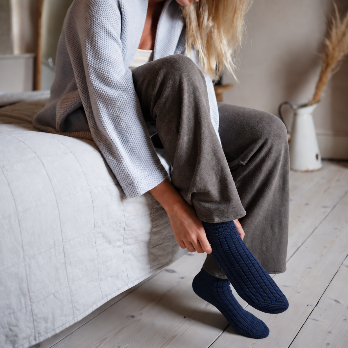 A woman sitting on a bed in a bedroom, wearing dark navy ribbed knit socks with her legs crossed.