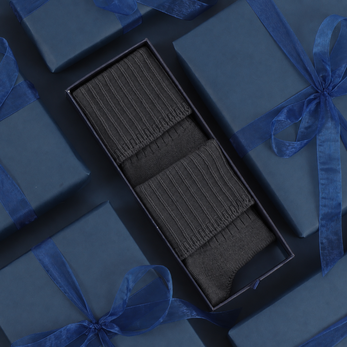 A pair of dark charcoal ribbed socks presented in an open black gift box, surrounded by dark blue wrapped gift boxes tied with blue satin ribbons.
