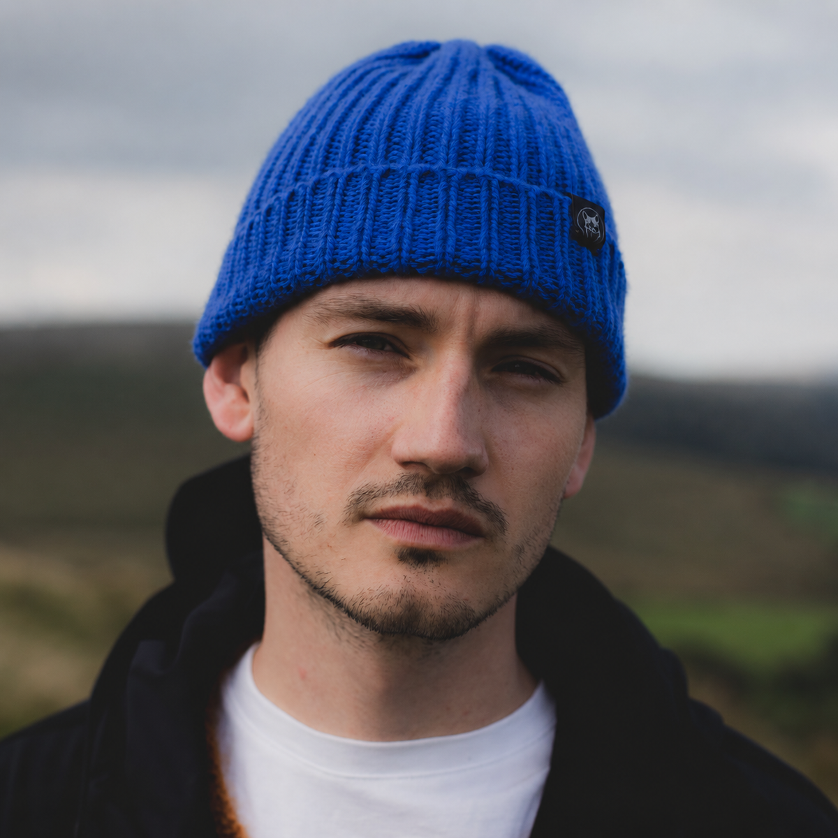 A man wearing a bright blue ribbed knit beanie with a small black logo patch on the side, photographed outdoors against a soft-focus hazy landscape.