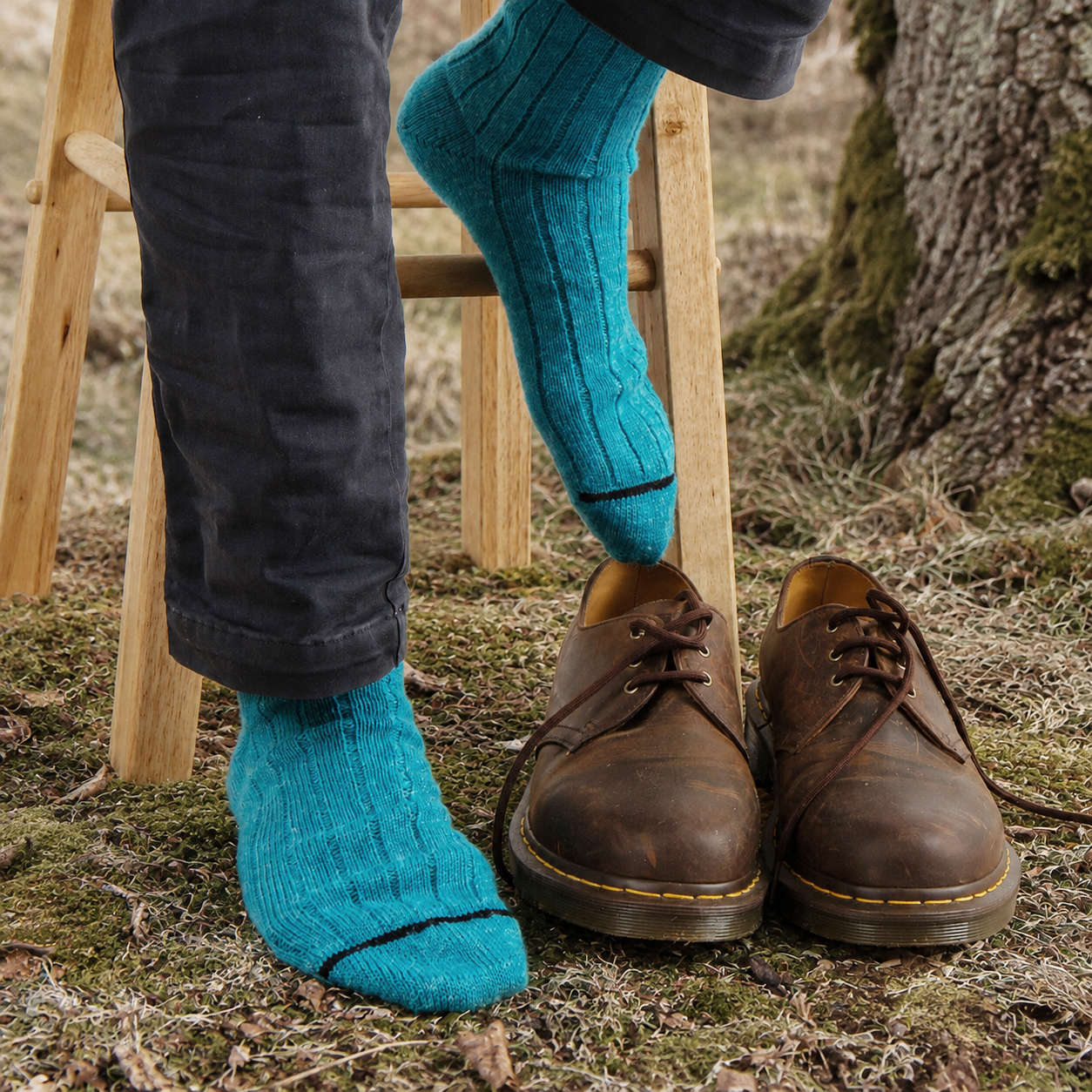 A person sitting outdoors on a wooden stool, wearing bright turquoise blue socks and dark charcoal pants, with a pair of brown leather lace-up shoes on the ground nearby.