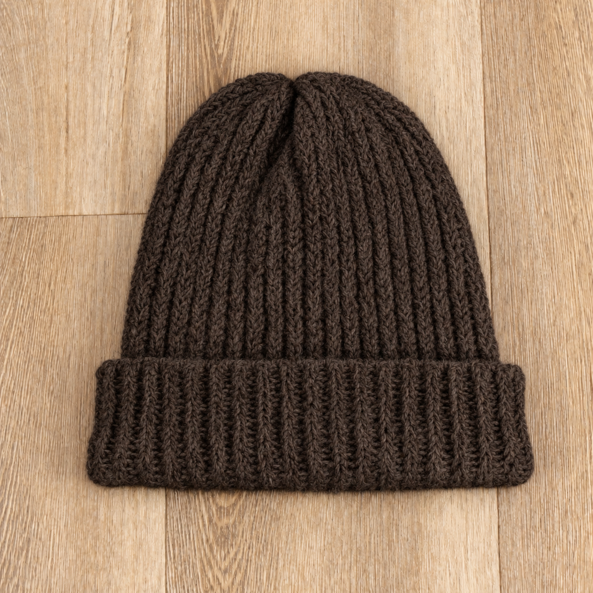 A dark brown ribbed knit beanie hat with a folded cuff, laid flat on a light wood plank background.
