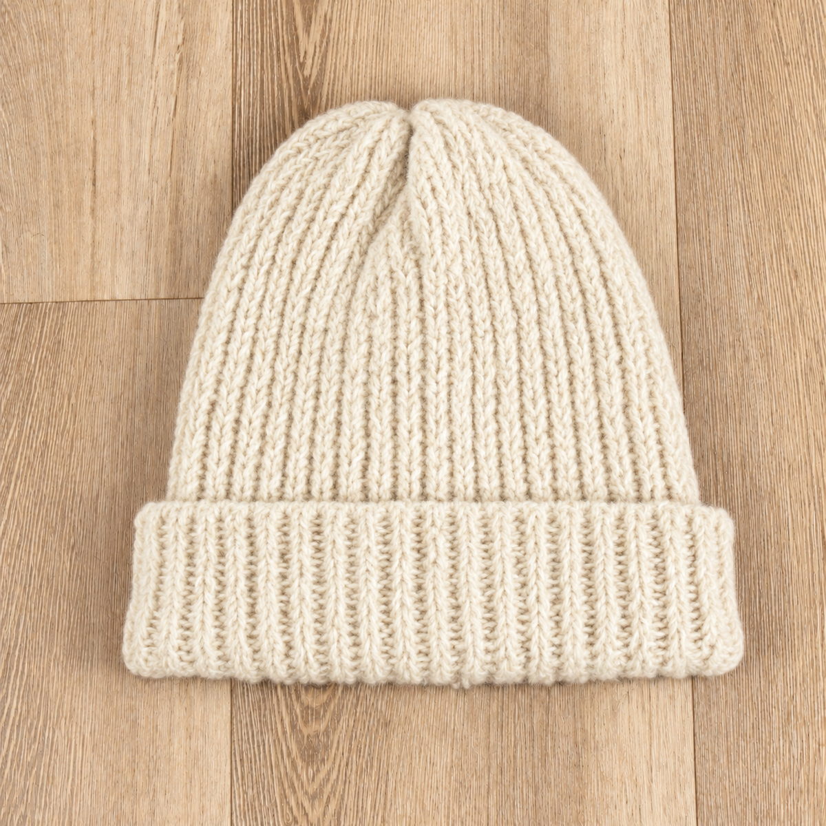 A cream-colored ribbed knit beanie hat laid flat on a light brown wooden floor.