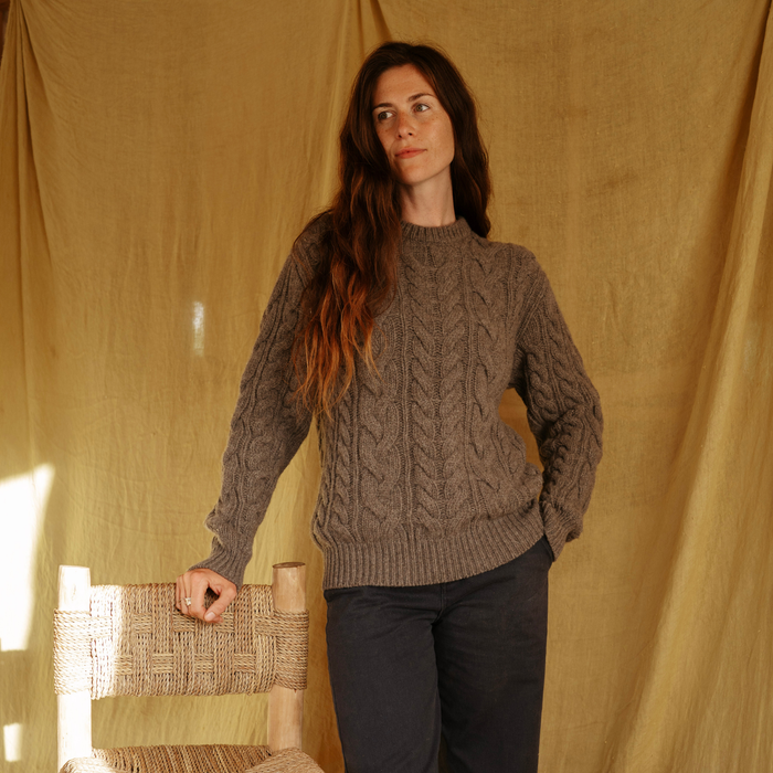 Woman wearing a brown cable knit sweater standing against a beige curtain.
