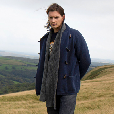 Man wearing a blue coat and grey scarf standing on a grassy hill with a scenic background