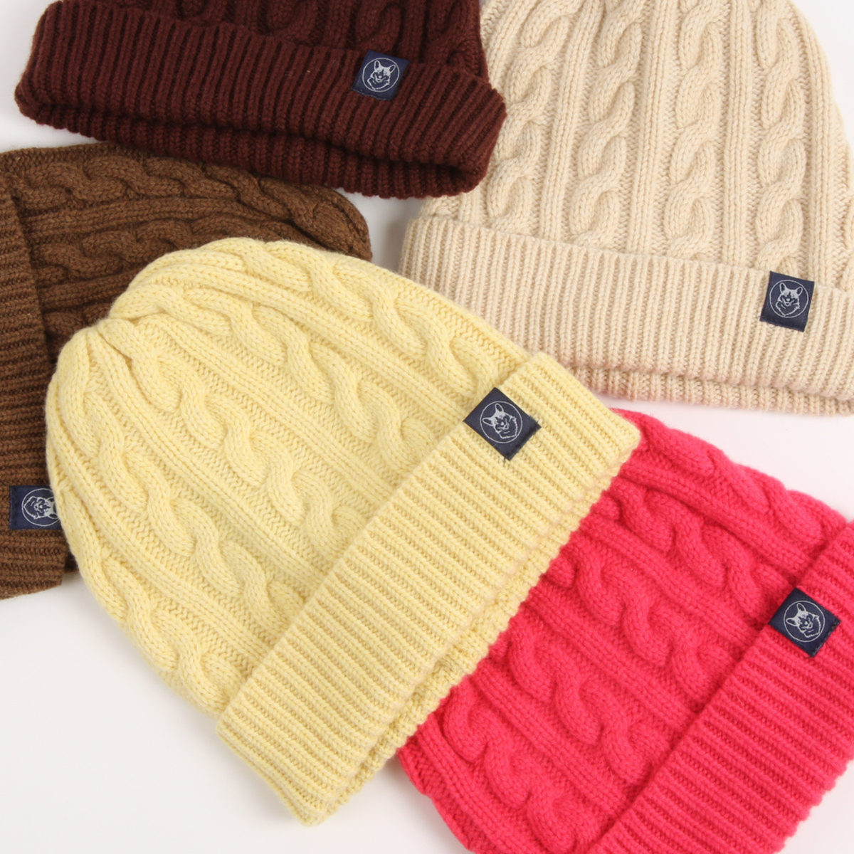 Four cable-knit beanie hats arranged on a white background, each with a small dark brand patch on the cuff. The hats are in dark brown, cream, pale yellow, and bright pink.