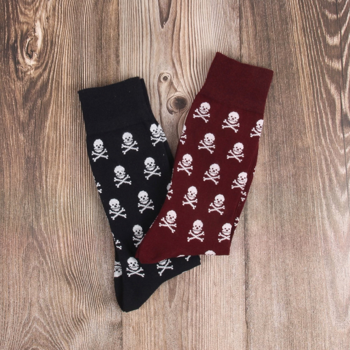 Two pairs of patterned crew socks laid flat on a wood surface: one navy pair with white skull and crossbones, and one burgundy pair with the same white skull and crossbones pattern.