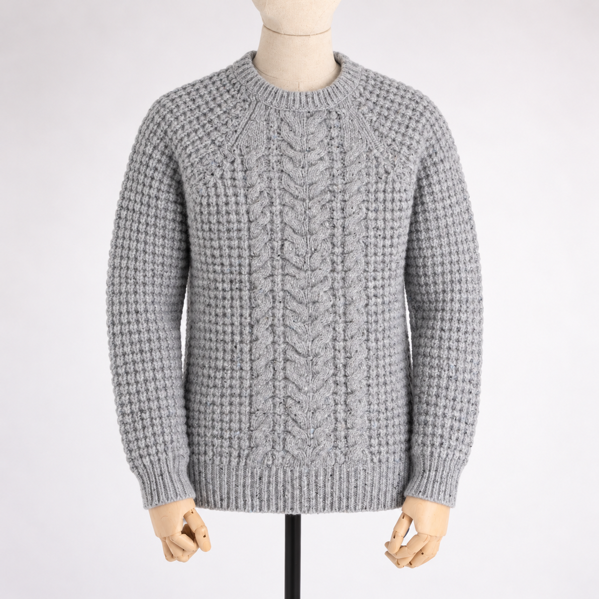 A light gray cable-knit crewneck sweater displayed on a mannequin torso with hands visible, set against a plain light gray background.
