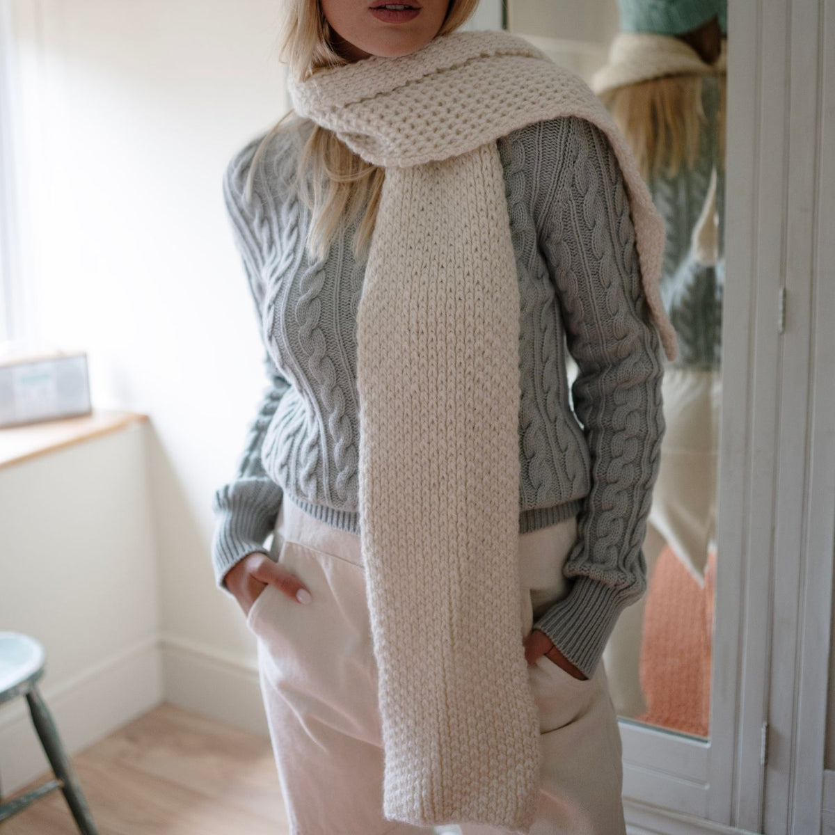 A woman wearing a aqua knitted sweater and a white cashmere scarf with a honeycomb pattern, standing in a room with a reflective surface.