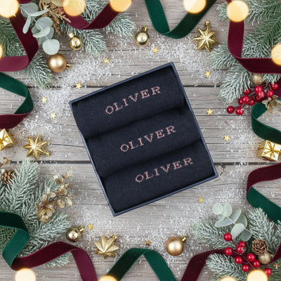 A set of three navy socks with the name 'OLIVER' personalized and knitted on the side, presented in a box on a wooden surface.