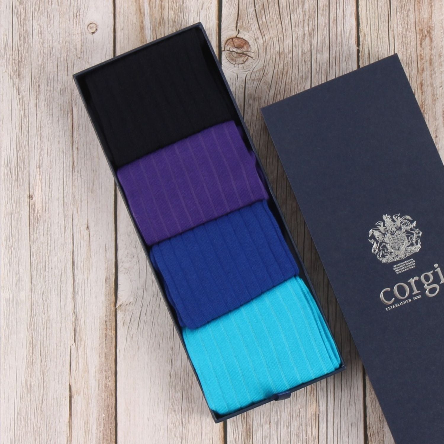 Three pairs of striped socks in purple, blue, and teal in an open black box on a wooden surface with 'Corgi' branding.