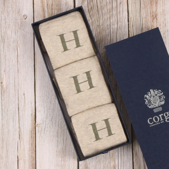 Three beige socks with 'H' monogram in a black box on a wooden surface, next to a navy blue box with a logo.