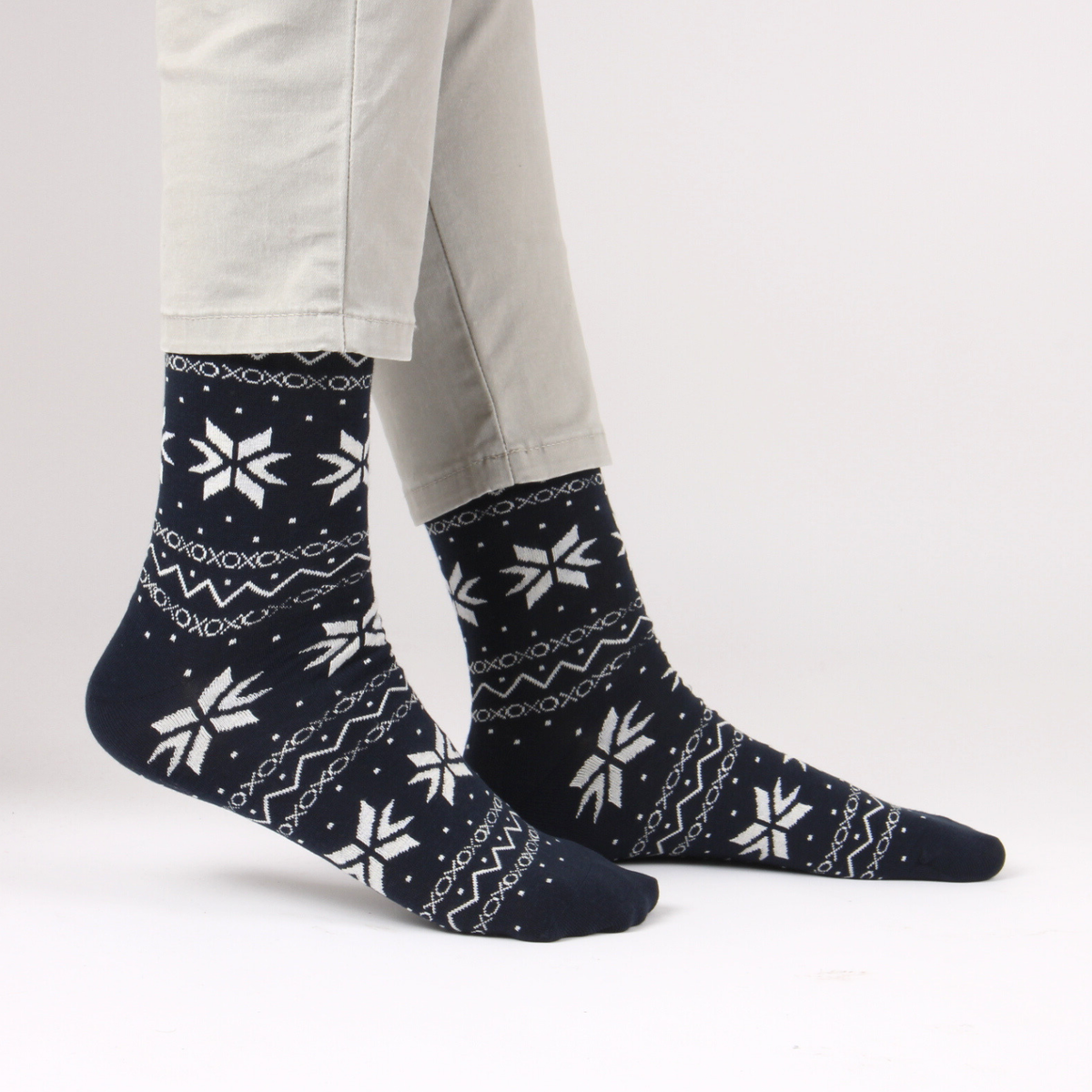 A pair of navy blue crew socks with a white Nordic/Fair Isle pattern of snowflakes, stars, and geometric lines, shown on a person's feet wearing light gray pants against a white studio background.
